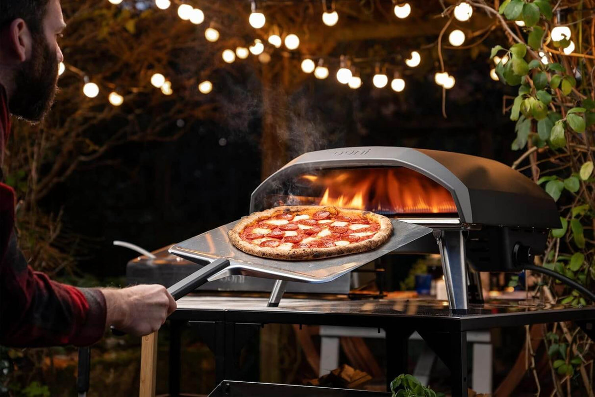 Outdoor pizza oven with Ooni Pizza Peel holding a pepperoni pizza, illuminated by string lights in a backyard setting.
