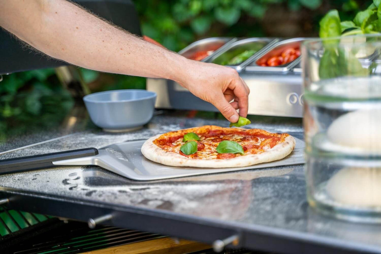 Person using Ooni Pizza Peel to place basil on freshly made pizza in outdoor kitchen setting.
