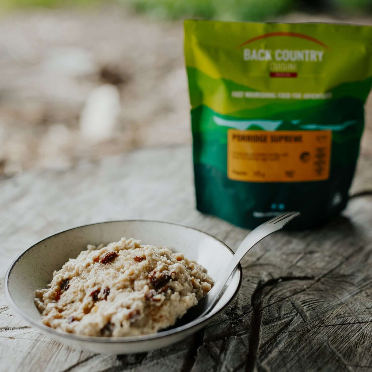 Back Country Cuisine Porridge Supreme camping breakfast with oats and dried fruit in a bowl, pouch in background on a wooden surface.