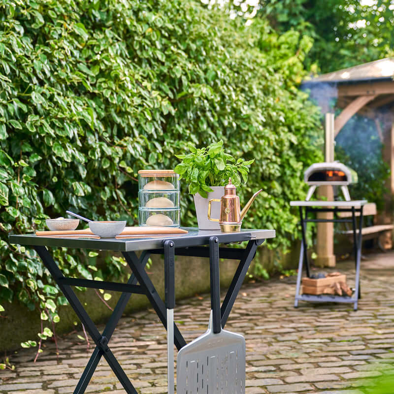 Ooni Folding Table setup with pizza prep essentials and an outdoor view, perfect for outdoor cooking.