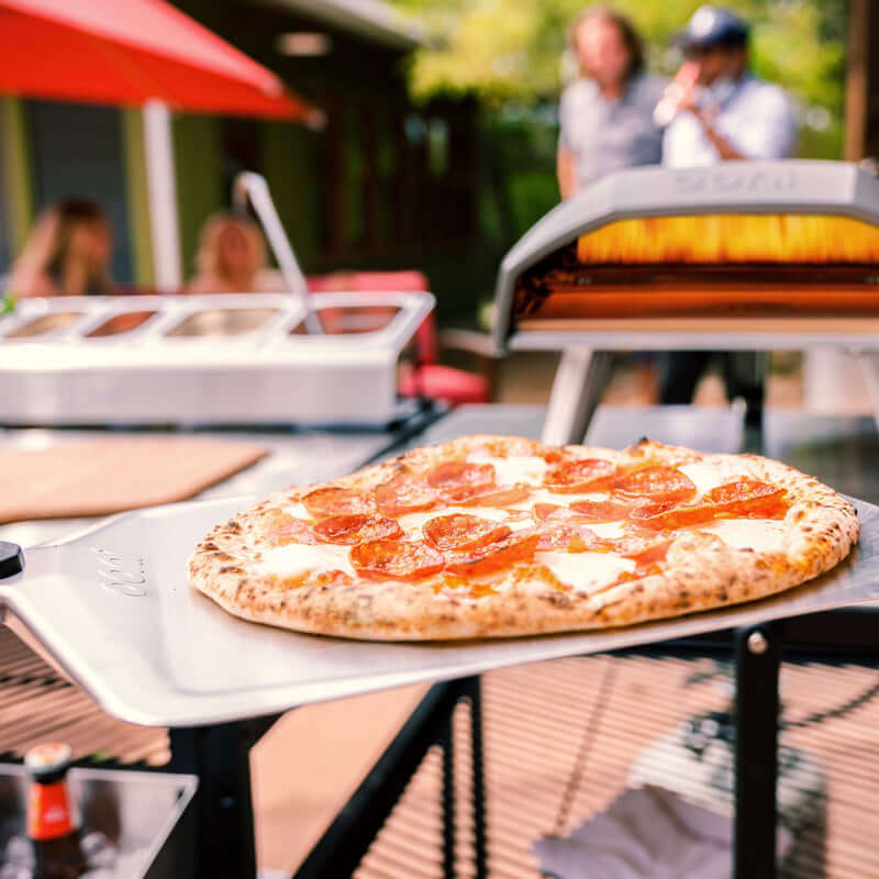 Ooni Pizza Peel in use with pepperoni pizza by outdoor oven, showcasing versatile cooking gear for backyard gatherings.