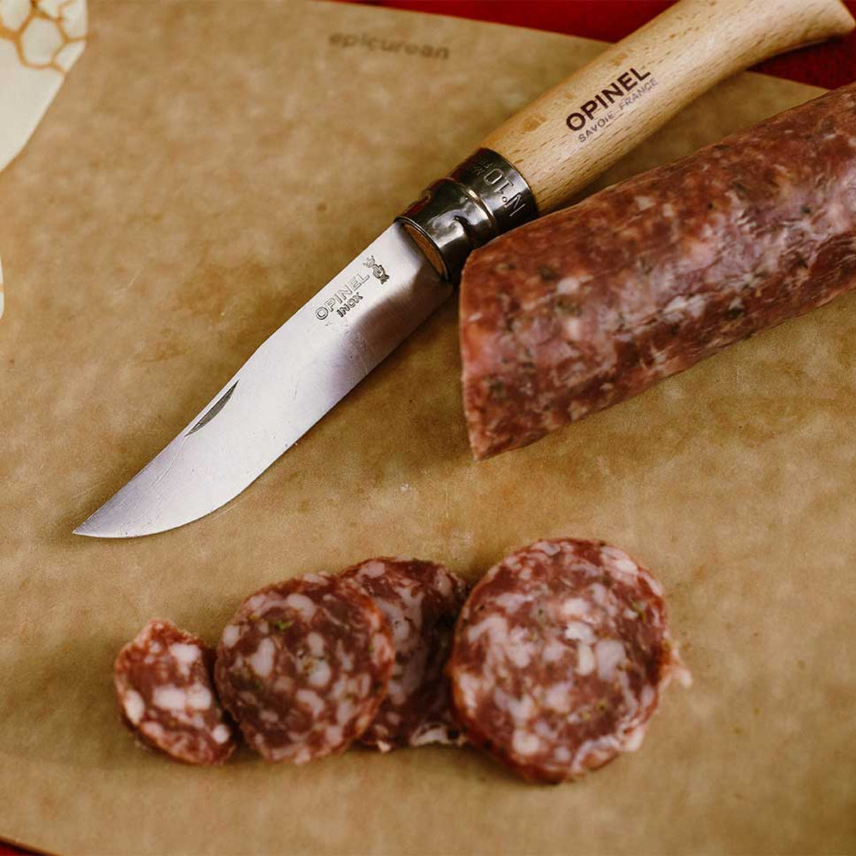 Opinel Traditional Stainless Steel Knife cutting salami on a wooden board, showcasing its sharp blade and wooden handle.