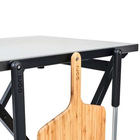 Ooni Folding Table with a stainless steel top and wooden pizza peel attached for easy access during pizza preparation.