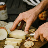 Using Opinel Traditional Stainless Steel Knife to slice cheese on a board for outdoor picnic with apple and crackers.