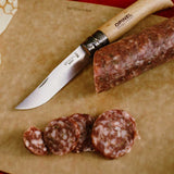 Opinel Traditional Stainless Steel Knife cutting salami on a wooden board, showcasing its sharp blade and wooden handle.
